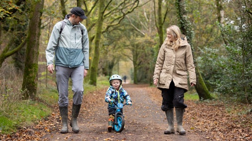 Young boy riding a balance bike along the path, with his mother and father walking either side of him